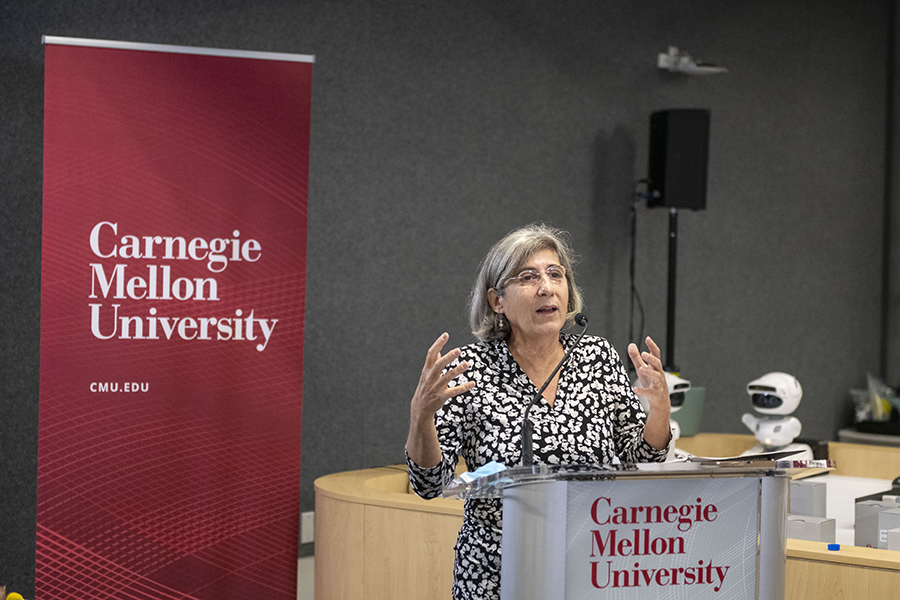 Manuela Veloso is shown behind a podium in a black and white dress, flanked by a CMU banner on the left and a small white robot on the right.